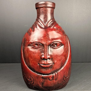 May include: A dark red, glossy ceramic vase featuring a sculpted human face on the front. The vessel has a rounded body, a narrow neck, and a smooth, polished finish with subtle dark marbling throughout the surface, creating a deep, rich color.