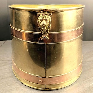May include: A cylindrical brass bucket featuring two horizontal copper bands and a prominent lion head ornament attached to the upper rim. The metal surface has a polished finish with visible seams and a metallic sheen throughout the structure.