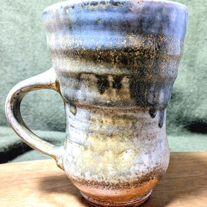 May include: Handmade ceramic mug with a unique, textured glaze. The mug features a cylindrical shape with a rounded base and a sturdy handle. The glaze transitions from blue at the top to brown and white, creating a rustic, earthy aesthetic. A perfect addition to any kitchen.