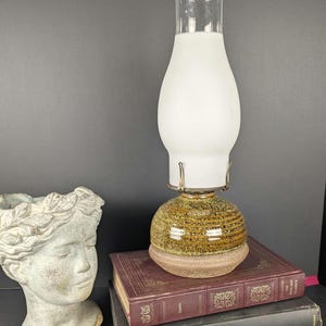 May include: A vintage table lamp with a frosted glass shade and a ceramic base. The base is a mix of brown and gold tones, and the lamp sits on a stack of books. The lamp has a clear glass chimney.