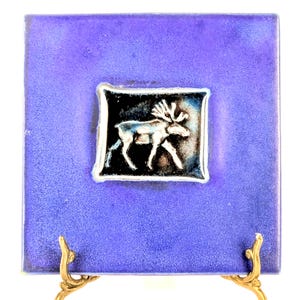 May include: Square ceramic tile with a textured, deep purple background. A central inset features a silver moose design on a black and blue background. The tile is displayed on a gold-colored stand.