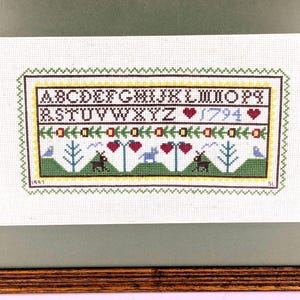May include: Framed cross-stitch sampler with a wooden frame. The sampler features the alphabet, the year 1794, and decorative elements like hearts, trees, and animals. The color palette includes green, brown, red, and blue.