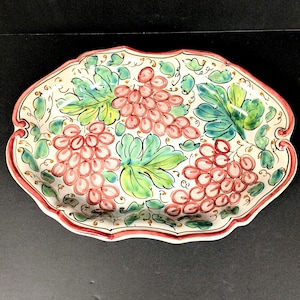 May include: Oval ceramic serving dish with a scalloped edge. The dish features a hand-painted design of pink grapes and green leaves on a cream background. The rim is outlined in red.