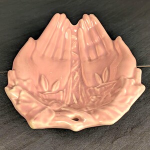 May include: A light pink ceramic trinket dish shaped like two cupped hands held together. The surface features a glossy finish with molded details of palms, fingers, and small leaf accents at the base. The dish has a small circular opening at the bottom.