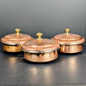 May include: Three copper-colored serving bowls with lids. Each bowl has a hammered texture, a silver-colored rim, and a gold-colored knob on the lid. The bowls are arranged on a dark surface.
