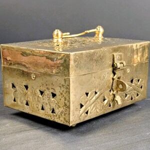 May include: A rectangular brass box with a hinged lid and a handle. The box features floral cutouts and decorative engravings. The latch is secured with a key. The box is a warm gold color and appears to be vintage.