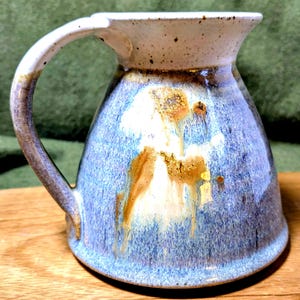 May include: Handmade ceramic mug with a unique glaze. The mug features a white rim and handle, with a blue and brown mottled design on the body. The mug has a curved handle and a wide opening. The mug is suitable for hot or cold beverages.