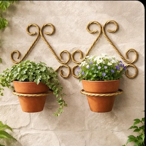 May include: Two terracotta pots filled with greenery and flowers hang from ornate, gold-colored metal wall planters. The planters have a decorative scroll design and are mounted on a textured, light-colored stone wall.