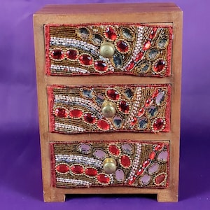 May include: A small wooden jewelry box with three drawers. Each drawer front is decorated with red, gold, and silver beads and gemstones. The drawer pulls are round and brass-colored. The box is made of light brown wood.