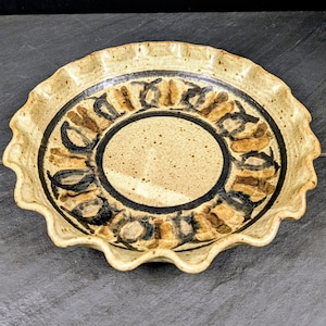 May include: A round ceramic pie dish with a ruffled, scalloped edge. The dish features a speckled beige glaze with a prominent dark brown circular band in the center and a ring of repeating dark brown and tan brushstroke patterns along the rim.