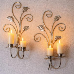 May include: Two silver-colored wall sconces, each with a floral and leaf design, hold two lit, ivory-colored candles. The sconces are mounted on a textured, off-white wall, creating a warm, inviting ambiance.