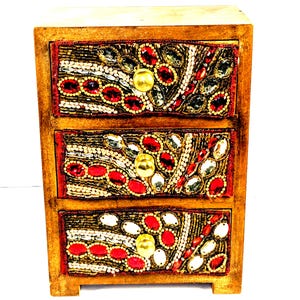 May include: A small, three-drawer wooden chest with ornate beaded embellishments. The drawers feature red, gold, white, and clear beads arranged in floral patterns. Each drawer has a round, gold-colored knob. The wooden frame has a light brown finish.