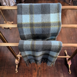 May include: A blue, gray, and yellow plaid blanket draped over a brass towel rack. The blanket has a fringed edge and a classic tartan pattern. The rack has ornate feet and is set against a dark wood floor.