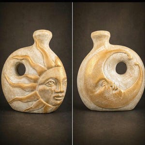 May include: Two-sided, cream-colored ceramic vase with a circular body and a narrow neck. One side features a golden sun face with radiating rays, and the other side depicts a golden crescent moon with a human face. The vase has a hole through the center.