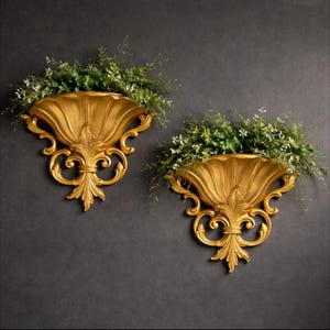 May include: Two ornate, gold-colored wall planters with a shell-like design. Each planter is filled with greenery and small white flowers. The planters have a detailed, scrollwork design and are mounted on a dark gray wall.