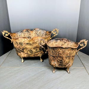 May include: Two metallic planters with a dark, weathered finish and intricate embossed leaf patterns. Each vessel features a scalloped rim, two ornate side handles, and three small feet. The larger planter is oval, while the smaller is square.