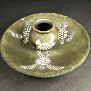 May include: A round, olive-green ceramic bowl featuring a central, smaller attached pot. The surface is decorated with three white, stylized sea turtle illustrations, with one turtle positioned on the central pot and two on the bowl base.