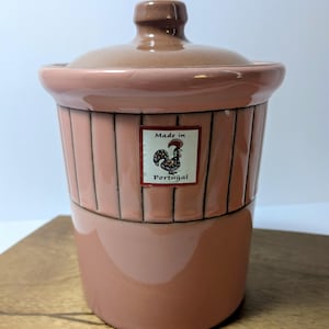 May include: A two-tone, pink ceramic jar with a lid. The jar has a cylindrical shape with vertical lines and a label that reads "Made in Portugal" with a rooster illustration. The lid and top of the jar are a darker shade of pink.