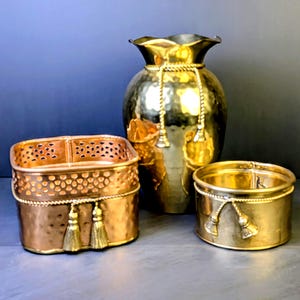 May include: A set of three gold-toned metal containers including a tall vase with a ruffled rim, a square basket with a perforated upper section, and a round bowl. Each piece features a decorative rope trim with hanging tassels in a metallic finish.