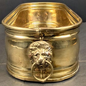 May include: A gold-colored metal container with a lion's head and ring handle. The container has a rectangular top and a cylindrical body. The lion's head is detailed, and the ring handle hangs below it. The container is made of a shiny, reflective metal.