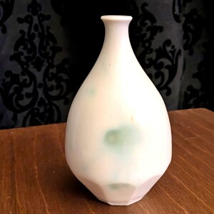 May include: A white ceramic vase with a rounded body and a narrow neck. The vase has a faceted base and a light green splotch on the front. The vase is displayed on a wooden surface.