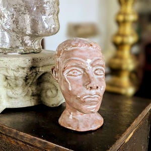 May include: A ceramic sculpture of a human head with a distressed, textured finish. The head is a light reddish-brown color with white accents around the eyes and mouth, and a tear-like detail. The sculpture is on a dark wooden surface.
