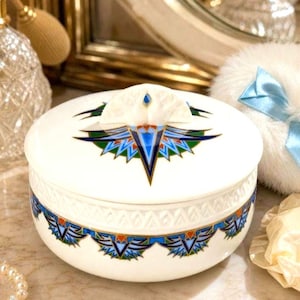 May include: A white ceramic trinket box with a lid featuring a geometric design in blue, green, and orange. The box has a matching decorative band around the base. A small, white, fan-shaped handle is on the lid.