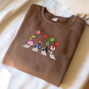 May include: A brown sweatshirt featuring a cartoon illustration of Princess Peach, Mario, Luigi, and Toad crossing a crosswalk, each holding a balloon. The design is a parody of the Beatles' Abbey Road album cover.