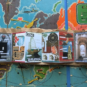 May include: A handmade travel-themed art journal with various images and text, including a sailboat, a bird, and architectural elements. The journal is open, revealing multiple pages with collaged designs and the word "Lose".