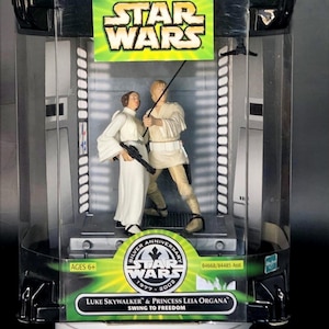 May include: A Star Wars collectible featuring Luke Skywalker and Princess Leia Organa figures. The figures are in a clear display case with a green and black Star Wars logo. The packaging includes the text "Silver Anniversary 1977-2002".