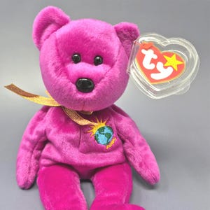 May include: A magenta plush bear with black eyes and nose, and a gold ribbon around its neck. The bear has an embroidered patch of the Earth with a sun design. A heart-shaped tag is attached.