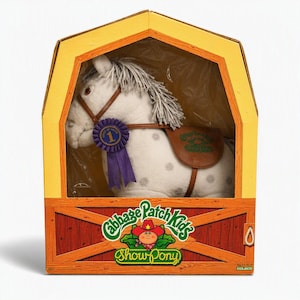 May include: A white plush toy horse with grey spots and a grey mane, wearing a brown bridle and saddle, inside a yellow barn-shaped box. The box features the text Cabbage Patch Kids Show Pony and a purple ribbon with the number 1 on the horse.