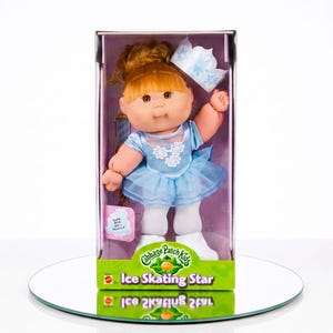 May include: A Cabbage Patch Kids Ice Skating Star doll with auburn hair in a bun, wearing a light blue dress with white floral accents, white tights, and white skates. The doll is packaged in a box with a blue crown and the text Ice Skating Star.
