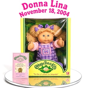May include: A Cabbage Patch Kids doll with blonde yarn hair in pigtails with purple plaid bows, wearing a pink long-sleeve shirt and purple plaid overalls. The doll is in a yellow box with the text Donna Lina November 18, 2004 and a birth certificate.