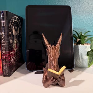 May include: A bronze-colored dragon figurine reading a book, with a tablet behind it. The dragon is holding a small, open book with yellow pages. The figurine is on a white surface, with a book and a plant in the background.