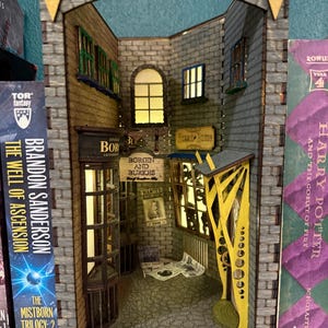 May include: A detailed diorama of a shop interior, featuring brick walls, arched doorways, and multiple windows. The shop has signs that read "BORGIN AND BURKES" and "WANTED". The diorama is flanked by book spines, including "The Well of Ascension" and "Harry Potter and the Goblet of Fire".