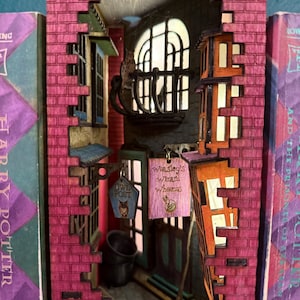 May include: A detailed diorama of a street scene, carved into a book. The scene features brick buildings in shades of pink and orange, with signs that read "Owl Post" and "Weasley's Wizard Wheezes". A broom and a bucket are also visible.
