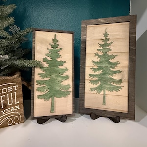 May include: Two wooden framed wall art pieces featuring a green Christmas tree design. The trees are cut out and mounted on a light wood background. The frames are dark brown and the art is displayed on a white surface.