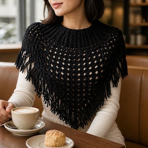 May include: A black crochet poncho featuring a textured, open-knit diamond pattern and a ribbed neckline. The garment is triangular in shape and finished with long, dangling fringe along the bottom edges, creating a layered, decorative appearance.