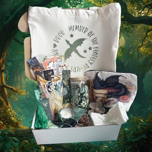 May include: A white tote bag and a white cosmetic bag with a dragon illustration are visible in an open box filled with fantasy-themed items. The tote bag has the text "Proud Member of the Fantasy Readers" and a dragon graphic. The box contains stickers, candles, and other accessories.