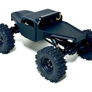 May include: A black remote-controlled truck with large, knobby tires. The truck has a flatbed and a light bar on top. The wheels are black, and the body is matte black. The vehicle is designed for off-road use.