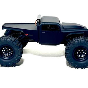 May include: A black, remote-controlled truck with large, knobby tires. The truck has a flatbed and a cab with a roll bar. The tires are designed for off-road use, and the vehicle is set against a white background.