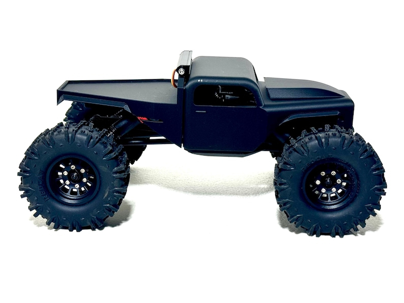 May include: A black, remote-controlled truck with large, knobby tires. The truck has a flatbed and a cab with a roll bar. The tires are designed for off-road use, and the vehicle is set against a white background.