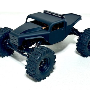 May include: A matte black remote control truck with large, knobby tires. The truck has a roll cage, a light bar, and a flatbed. The front grill has a mesh design. The vehicle is designed for off-road use.