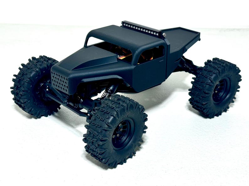 May include: A matte black remote control truck with large, knobby tires. The truck has a roll cage, a light bar, and a flatbed. The front grill has a mesh design. The vehicle is designed for off-road use.