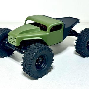 May include: A small, olive green model truck with large, black, off-road tires. The truck has a detailed front grille and a black bed. The tires are heavily treaded for traction. The vehicle is designed for off-road use.
