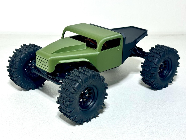 May include: A small, olive green model truck with large, black, off-road tires. The truck has a detailed front grille and a black bed. The tires are heavily treaded for traction. The vehicle is designed for off-road use.