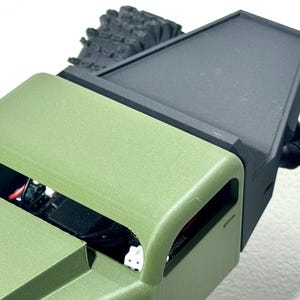 May include: Close-up of a green and black model vehicle. The vehicle has a green body with an open top, revealing internal components. The black section appears to be a cover or attachment. The tires are black and textured.