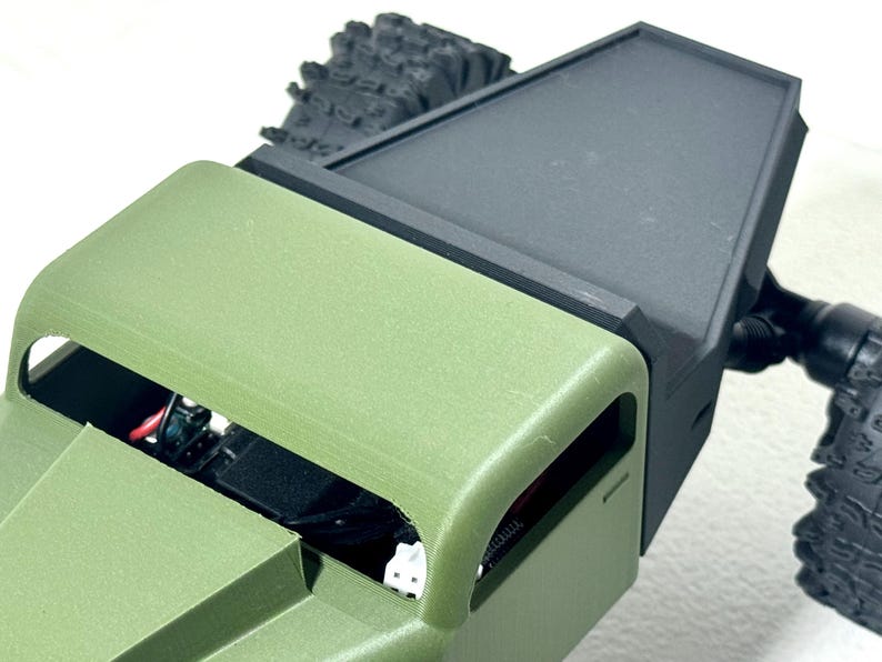 May include: Close-up of a green and black model vehicle. The vehicle has a green body with an open top, revealing internal components. The black section appears to be a cover or attachment. The tires are black and textured.