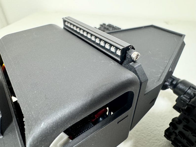 May include: A black, rectangular light bar with multiple small LED lights mounted on a black, angular structure. The structure appears to be part of a larger, possibly robotic, device. The image is a close-up.